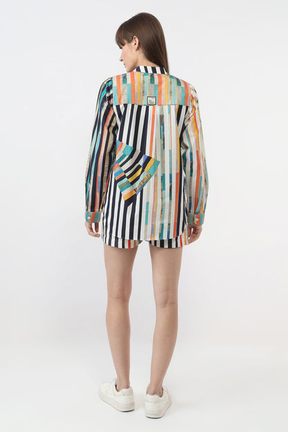 A model viewed from the back wearing a long-sleeve mini shirt with a colorful vertical stripe pattern in black, white, orange, blue, and yellow, paired with white sneakers on a light gray background.