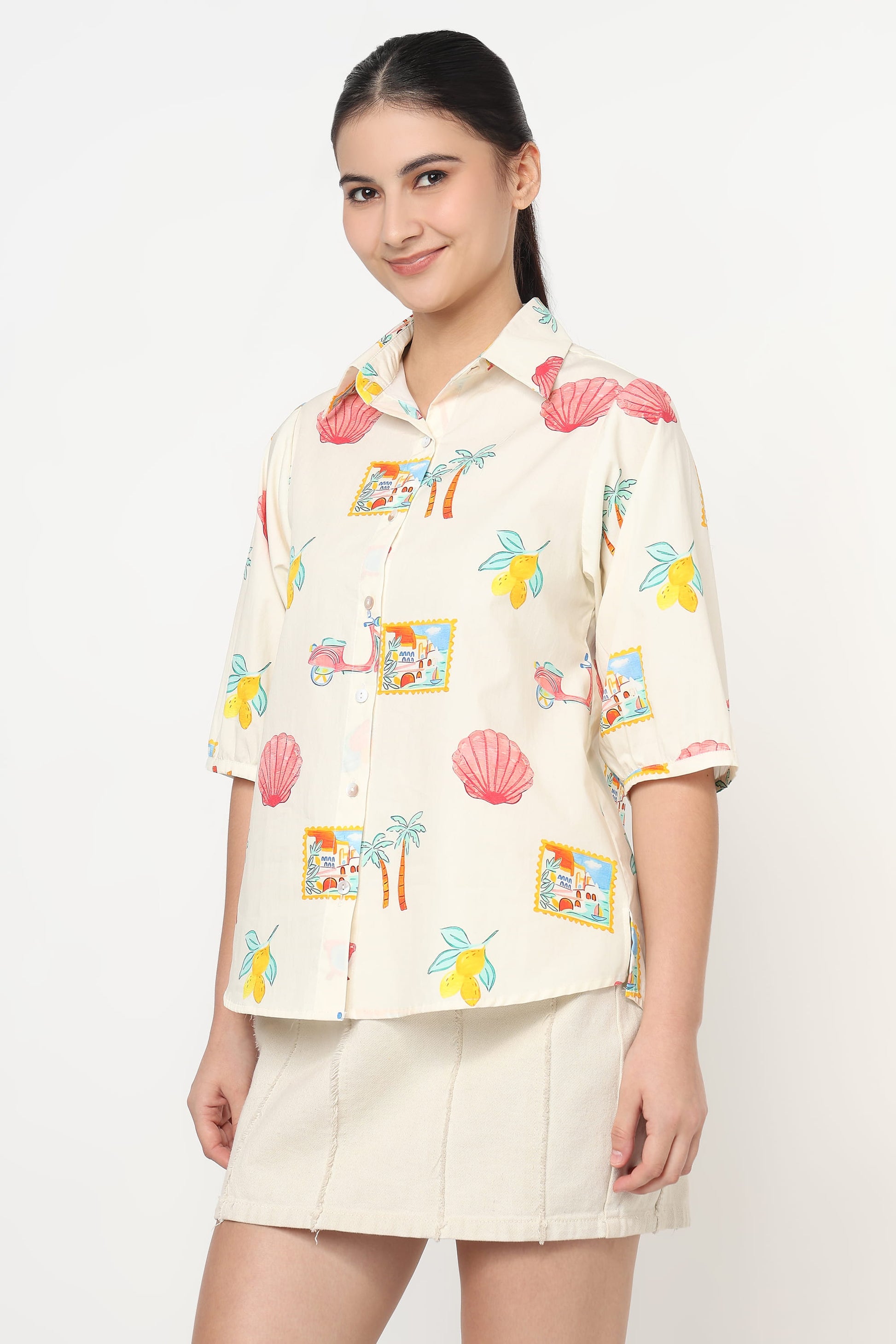 A woman modeling a cream-colored short-sleeve button-up shirt with a colorful tropical print, paired with a matching cream mini skirt, against a plain white studio background.