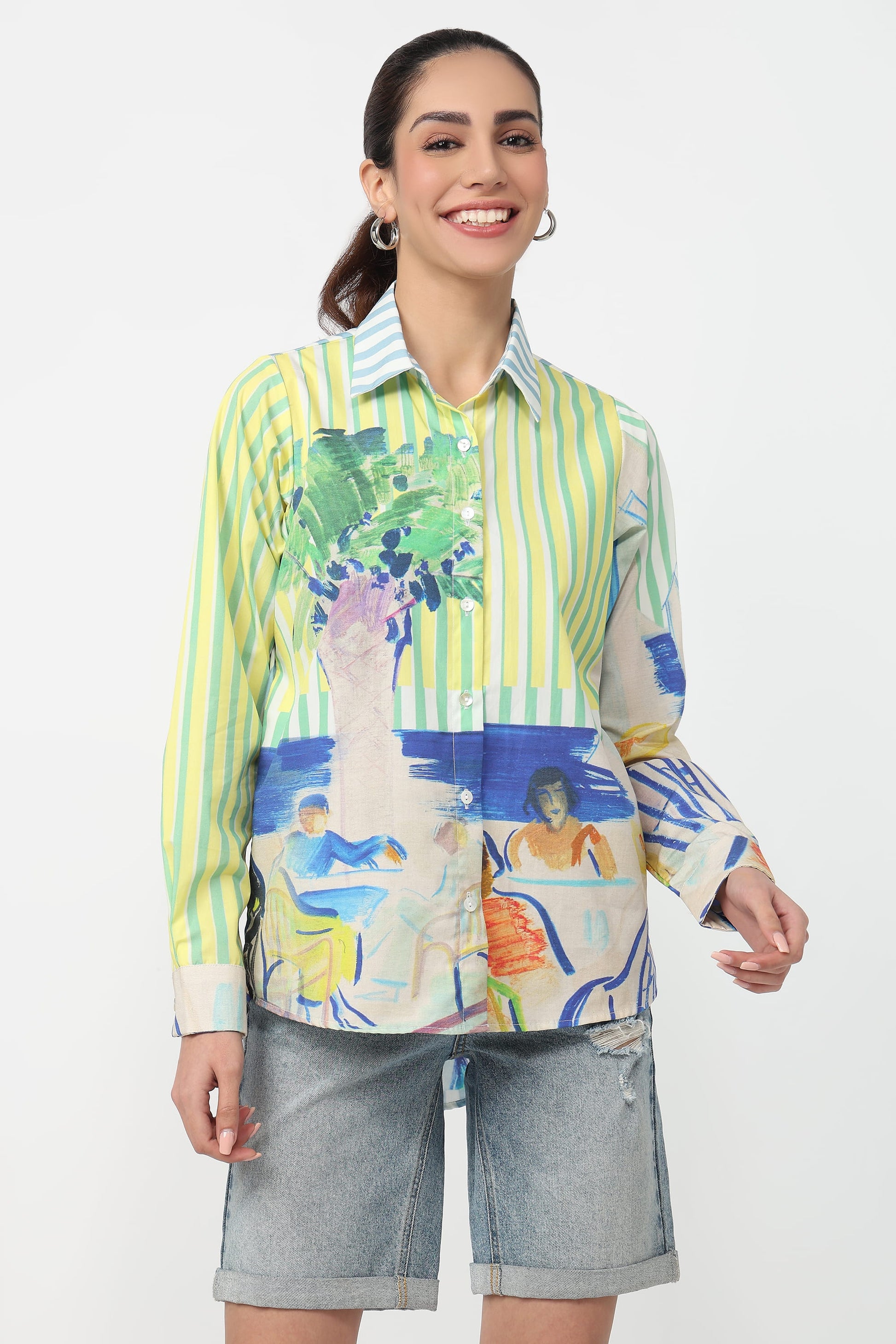 A woman modeling a long-sleeve button-up shirt with a colorful abstract print and vertical lime green and white stripes, paired with gray denim shorts against a plain white background.