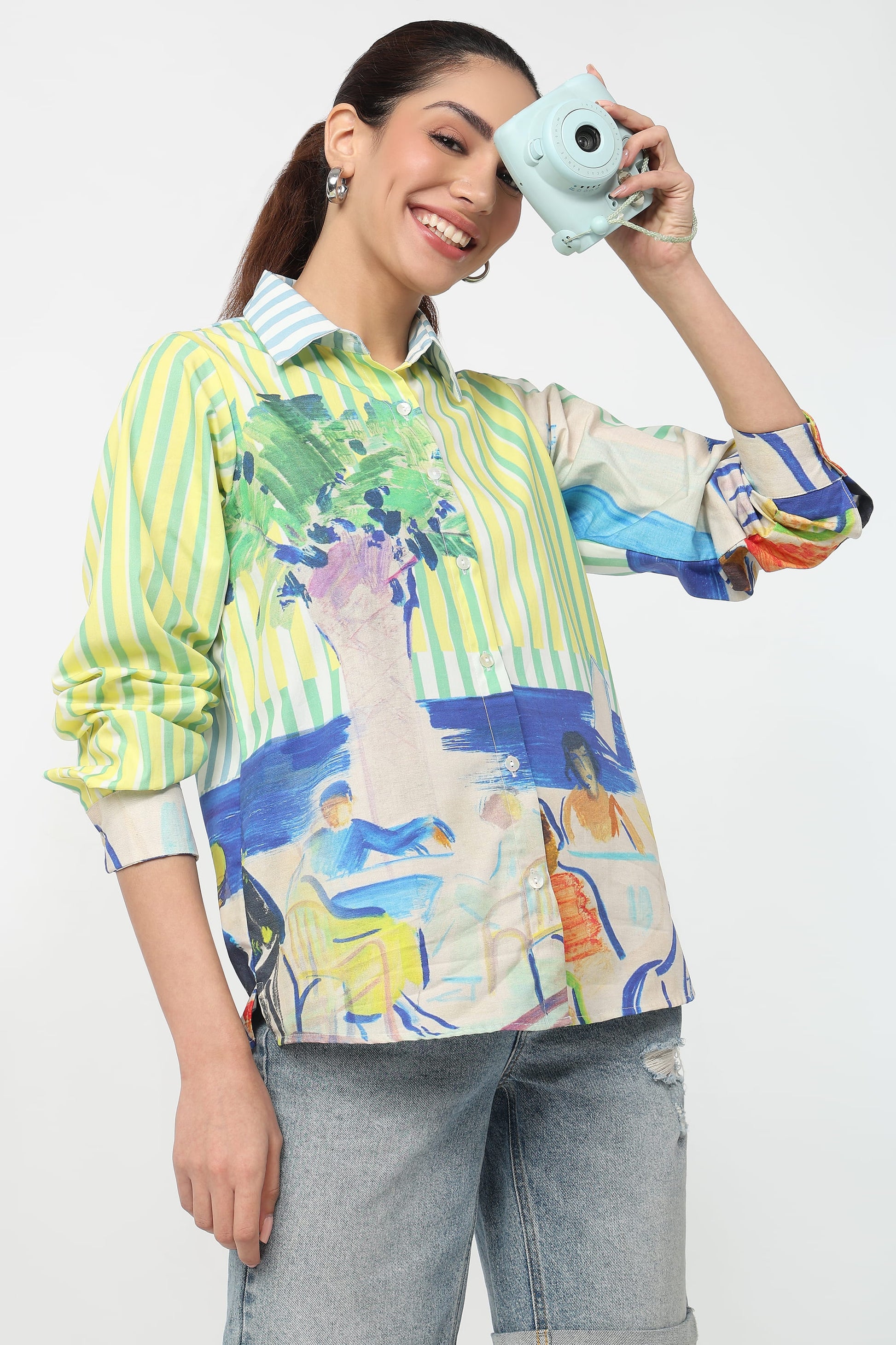 A woman wearing a colorful, long-sleeve button-up shirt with a mix of green and white vertical stripes and a blue, green, and yellow abstract landscape print. She is holding a light blue instant camera to her eye against a plain white background.