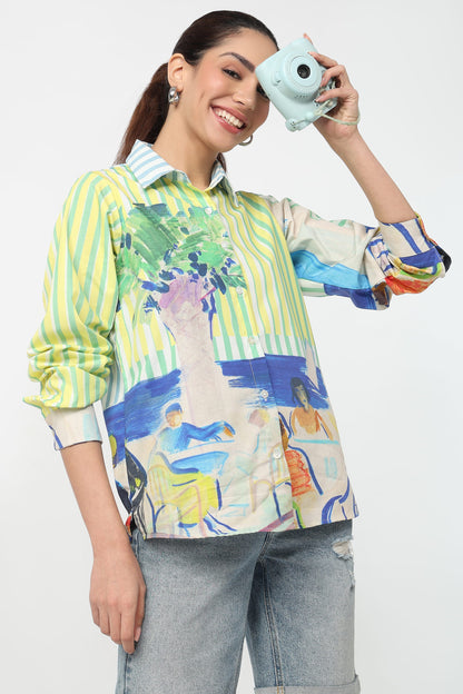 A woman wearing a colorful, long-sleeve button-up shirt with a mix of green and white vertical stripes and a blue, green, and yellow abstract landscape print. She is holding a light blue instant camera to her eye against a plain white background.