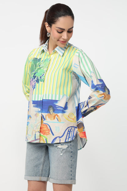 A woman wearing a long-sleeve button-up shirt with a vibrant multicolor print featuring green and yellow vertical stripes on one side and a beach-themed illustration with blue, orange, and white elements on the other, paired with light-wash denim shorts against a plain white background.