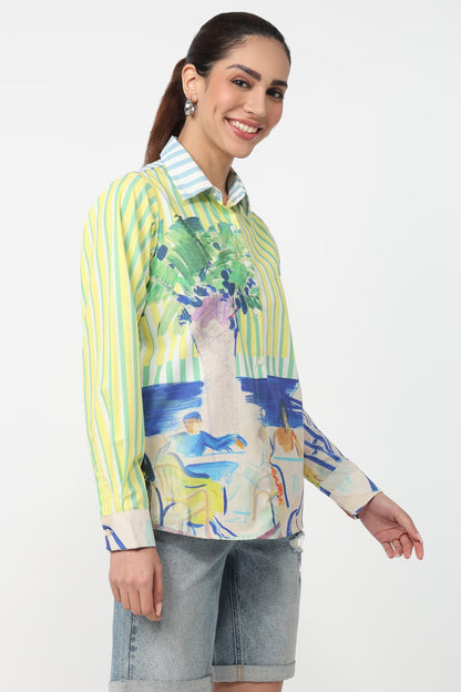 A woman modeling a long-sleeve button-up shirt with a vibrant green, yellow, blue, and white abstract landscape print, paired with light-wash denim shorts against a plain white background.