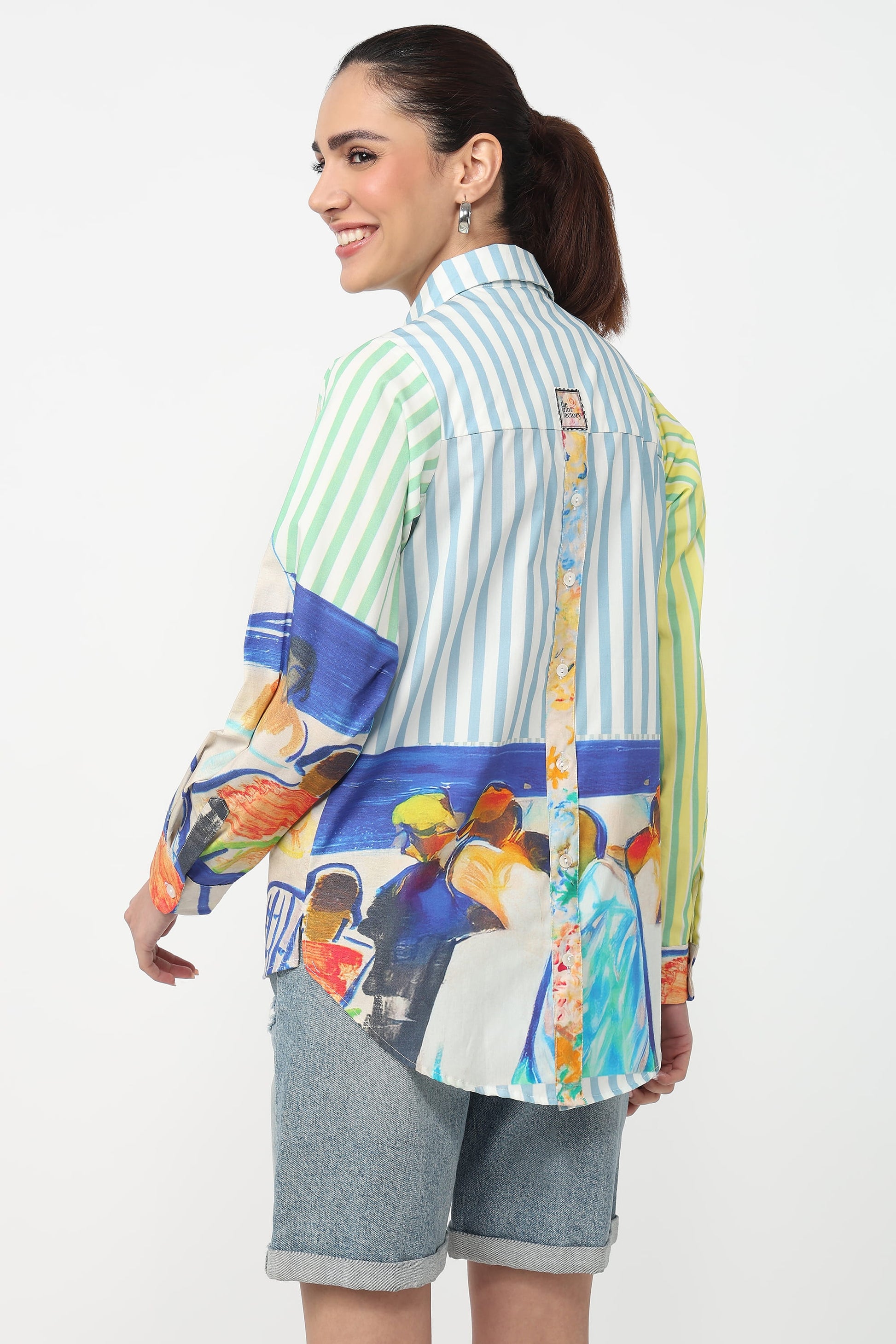 A woman shown from the back wearing a long-sleeve button-up shirt with a multicolor abstract print and striped panels, paired with denim shorts, against a plain white studio background.