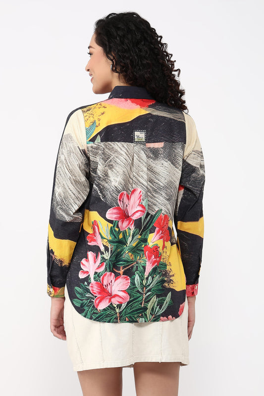 A woman shown from the back wearing a long-sleeve patchwork button-up shirt with a bold abstract print and large pink hibiscus flowers, paired with a cream mini skirt, against a plain white studio background.