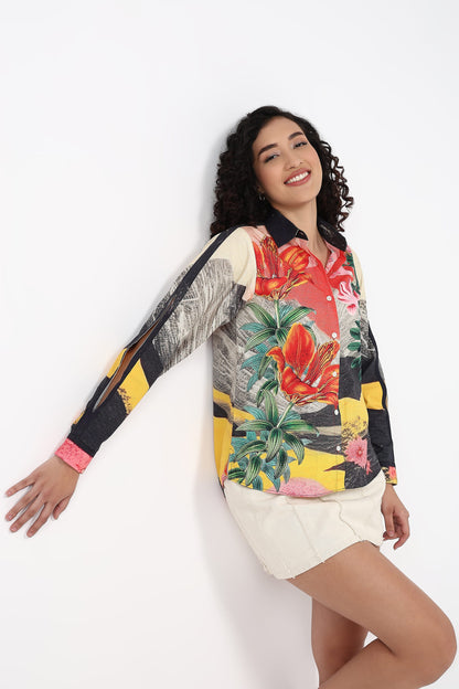 A woman modeling a long-sleeve collared shirt with a vibrant tropical floral print, featuring red hibiscus flowers, green palm leaves, and yellow accents, paired with off-white shorts against a plain white background.