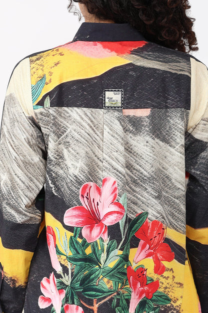 Close-up back view of a person wearing a long-sleeve collared shirt with a bold abstract and floral print. The shirt features black, gray, yellow, red, pink, and green patterns, with a small sewn label near the upper back.