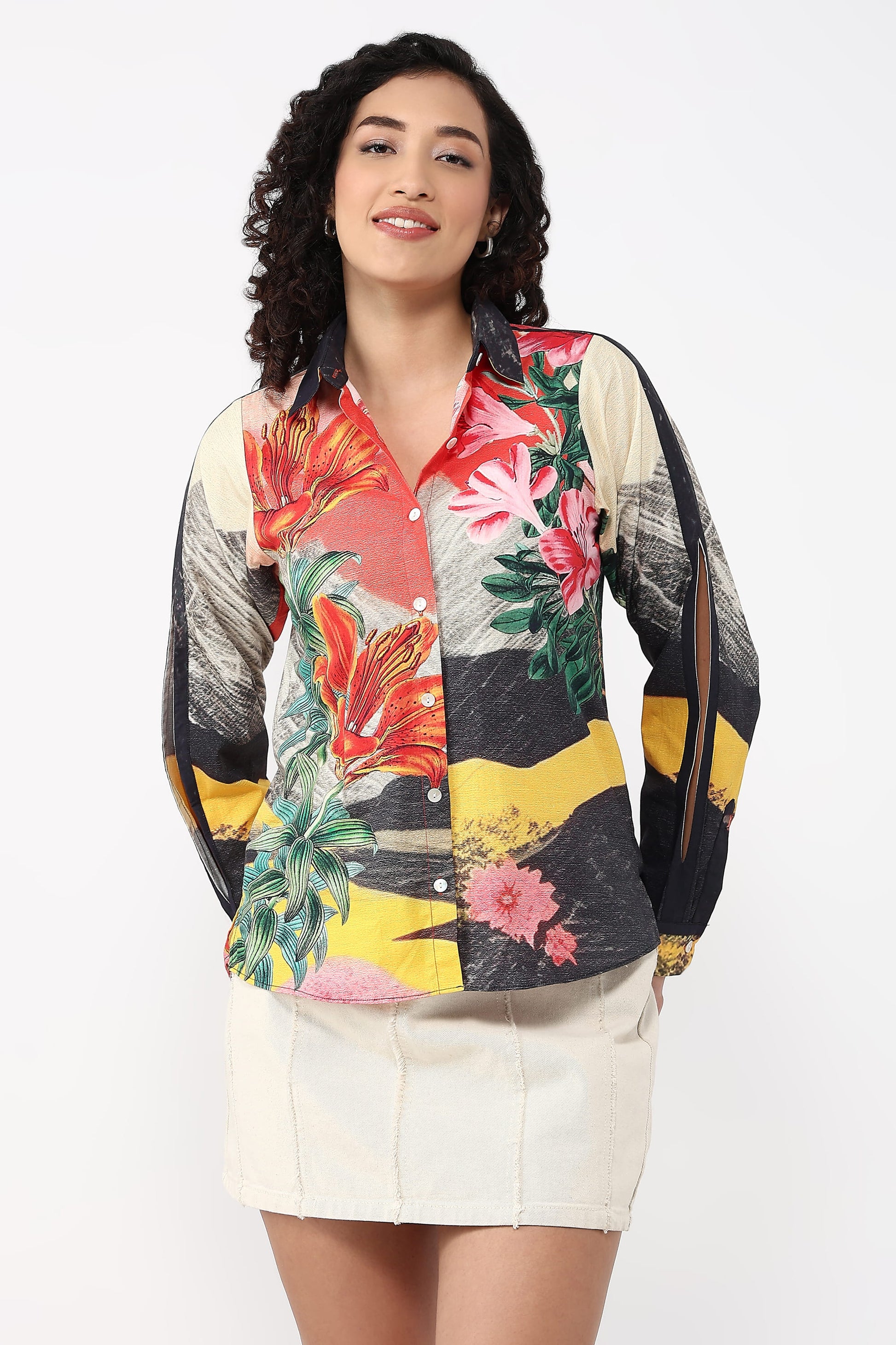 A woman modeling a long-sleeve button-up shirt with a vibrant multicolor tropical and abstract print, paired with a cream-colored mini skirt, against a plain white studio background.