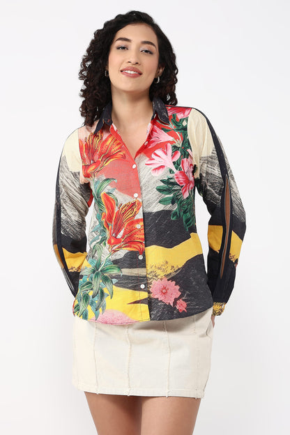 A woman modeling a long-sleeve button-up shirt with a vibrant multicolor tropical and abstract print, paired with a cream-colored mini skirt, against a plain white studio background.