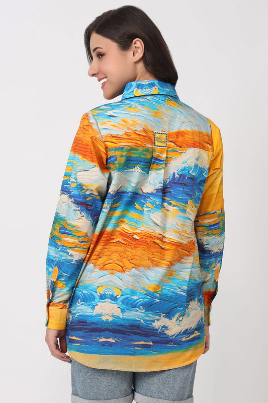 A woman shown from the back wearing a long-sleeve button-up shirt with a colorful abstract brushstroke pattern in blue, orange, yellow, and white, paired with light-wash denim jeans against a plain light gray studio background.