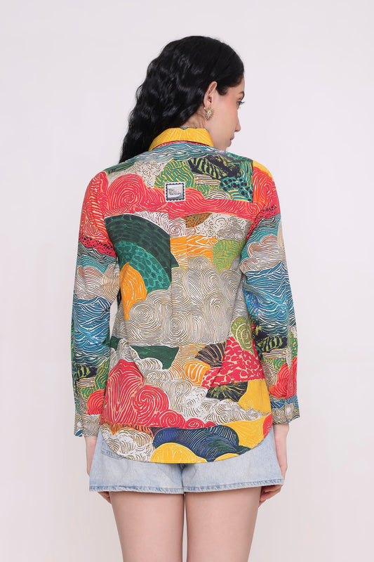 A woman viewed from behind wearing a long-sleeve button-up shirt with a vibrant, multicolored abstract pattern in red, yellow, green, blue, and white, paired with light-wash denim shorts against a plain light background.