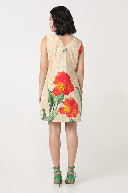 A woman viewed from the back wearing a sleeveless beige dress with a large red and orange floral print and green leaves, paired with green high-heeled sandals, against a light grey studio background.