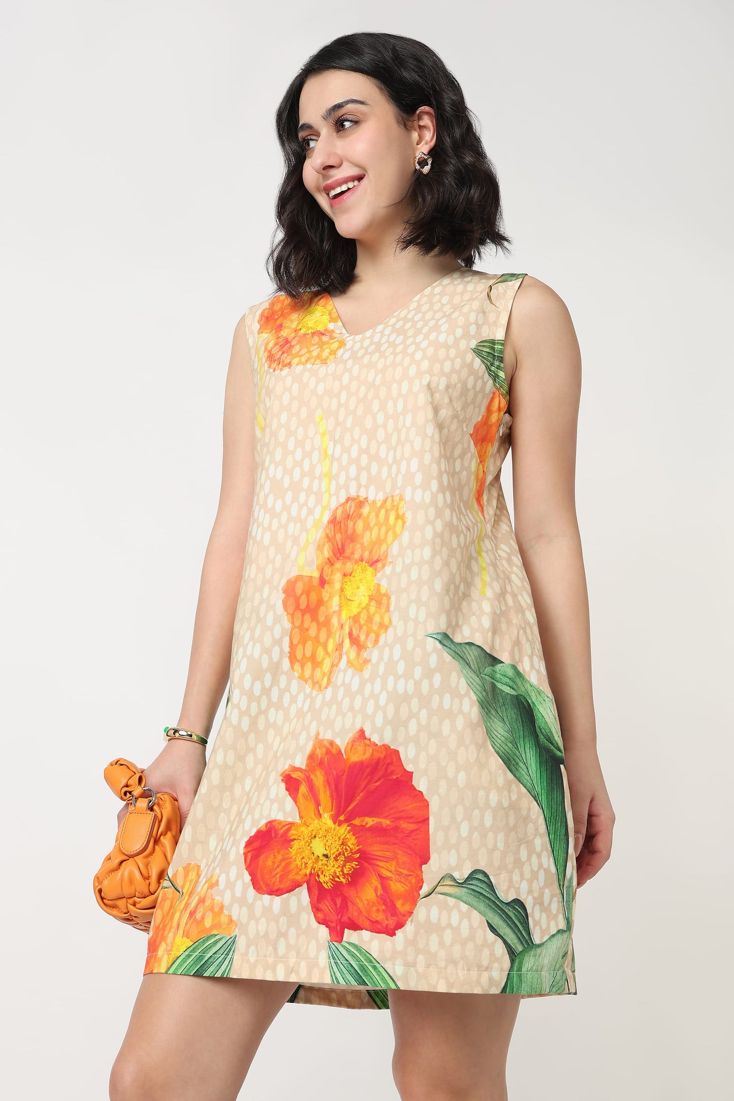 A woman wearing a sleeveless beige mini dress with a white polka dot pattern and large orange and red hibiscus floral prints, holding a textured orange clutch bag against a plain light grey background.