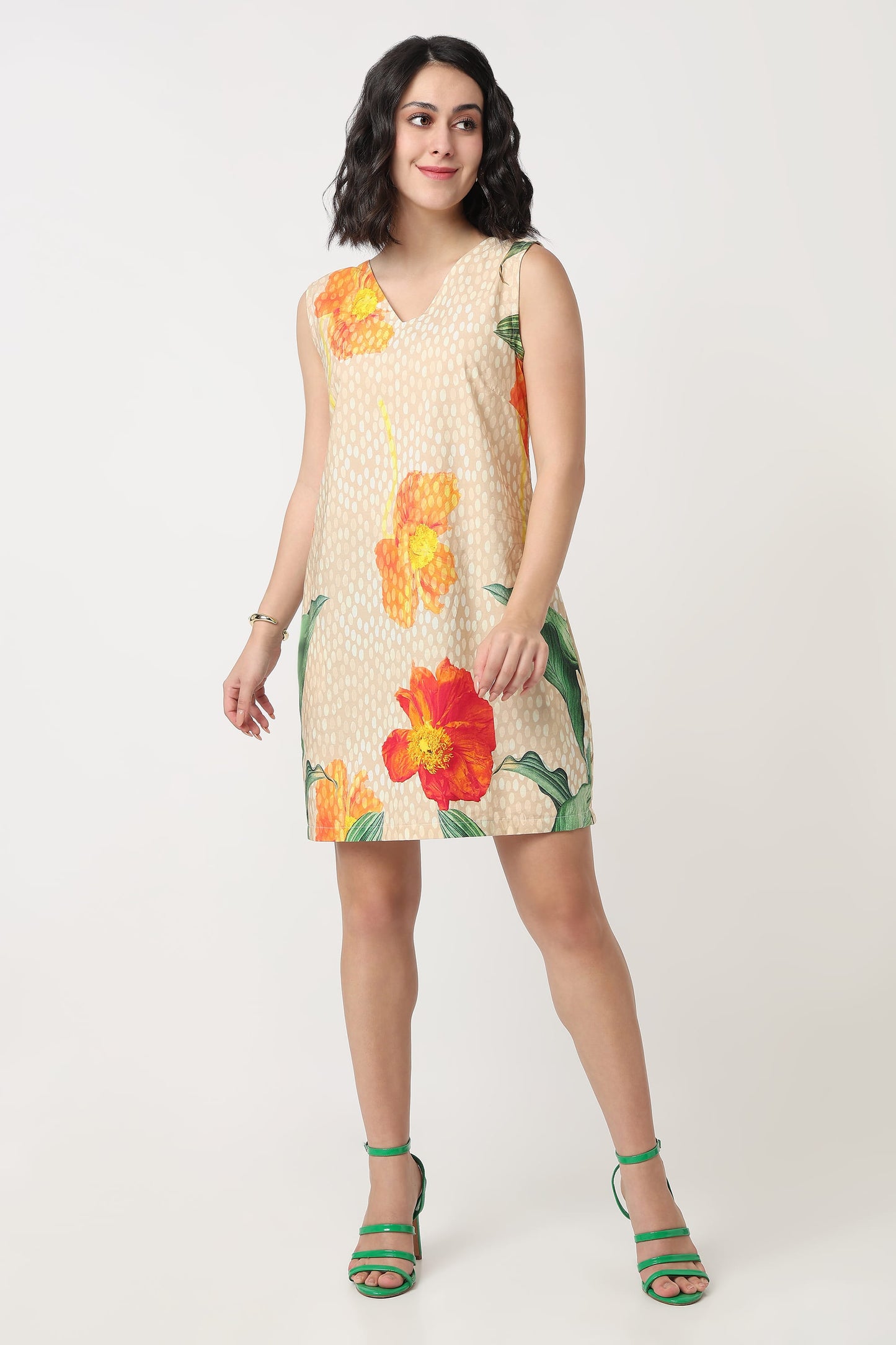 A woman wearing a sleeveless beige mini dress with a white polka dot pattern and large orange and red hibiscus floral prints, holding a textured orange clutch bag against a plain light grey background.