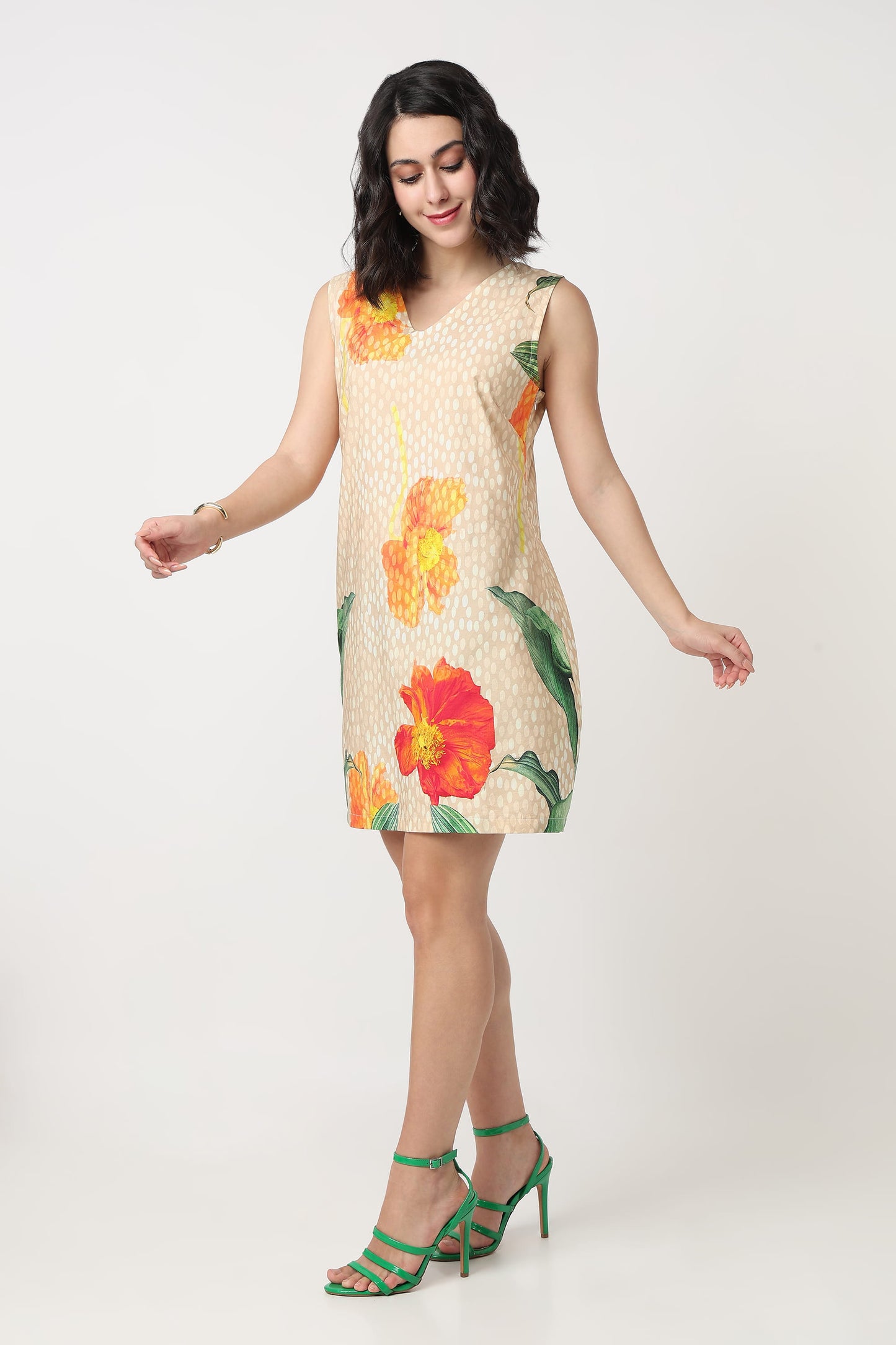 A woman wearing a sleeveless beige mini dress with a white polka dot pattern and large orange and red hibiscus floral prints, holding a textured orange clutch bag against a plain light grey background.