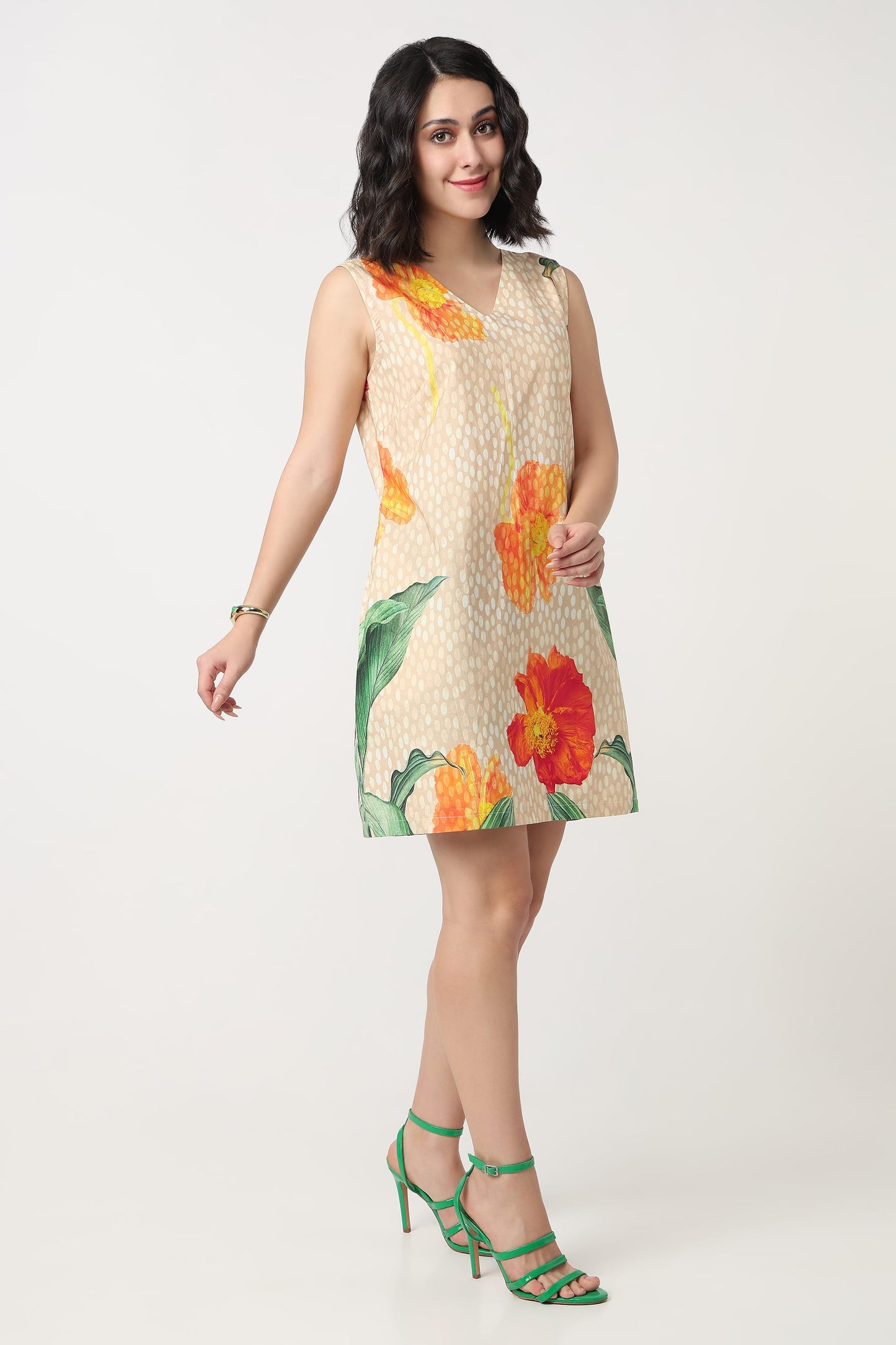 A woman wearing a sleeveless beige mini dress with a white polka dot pattern and large orange and red hibiscus floral prints, holding a textured orange clutch bag against a plain light grey background.
