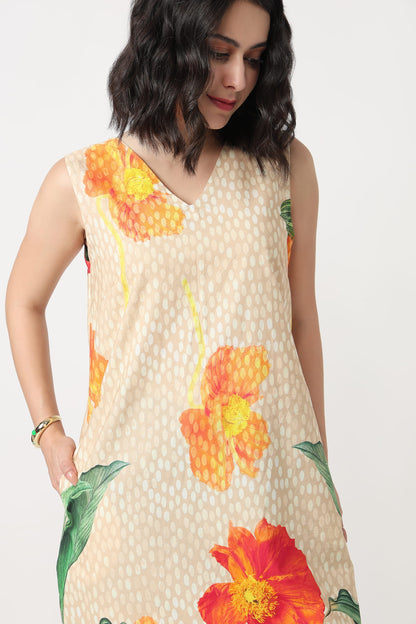A woman wearing a sleeveless beige mini dress with a white polka dot pattern and large orange and red hibiscus floral prints, holding a textured orange clutch bag against a plain light grey background.