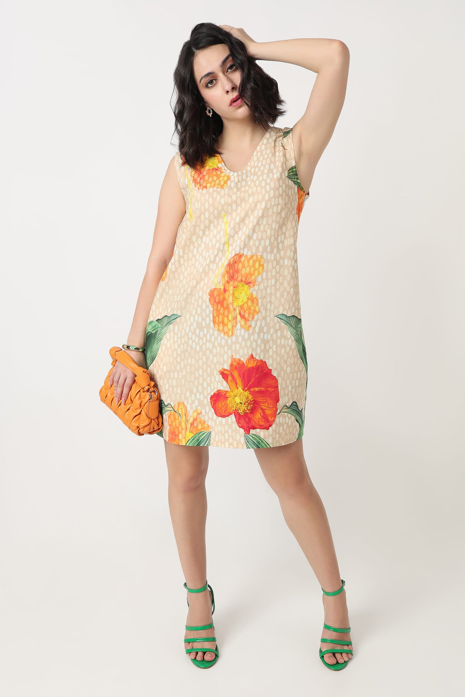 A woman wearing a sleeveless beige mini dress with a white polka dot pattern and large orange and red hibiscus floral prints, holding a textured orange clutch bag against a plain light grey background.