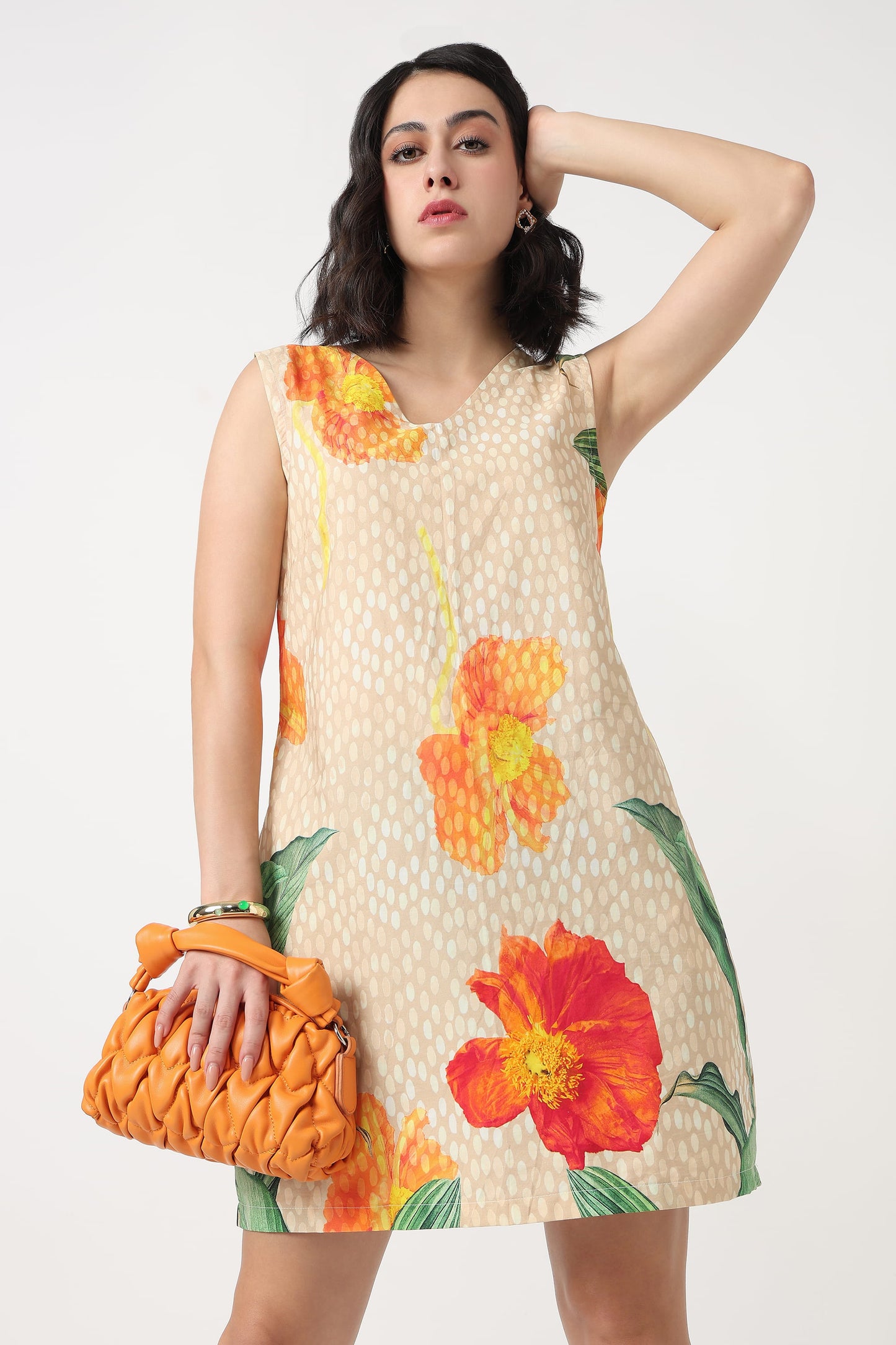 A woman wearing a sleeveless beige mini dress with a white polka dot pattern and large orange and red hibiscus floral prints, holding a textured orange clutch bag against a plain light grey background.