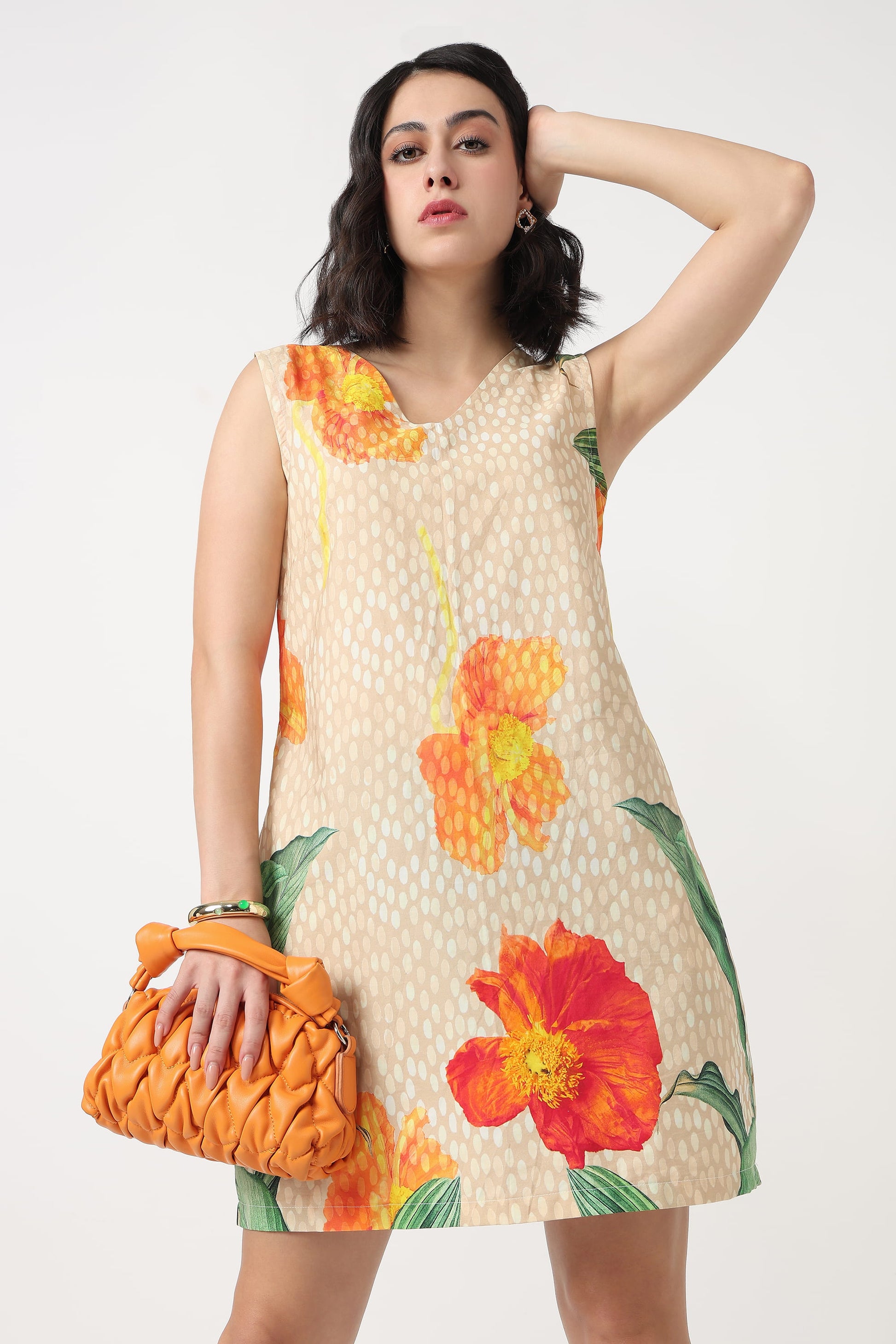 A woman wearing a sleeveless beige mini dress with a white polka dot pattern and large orange and red hibiscus floral prints, holding a textured orange clutch bag against a plain light grey background.