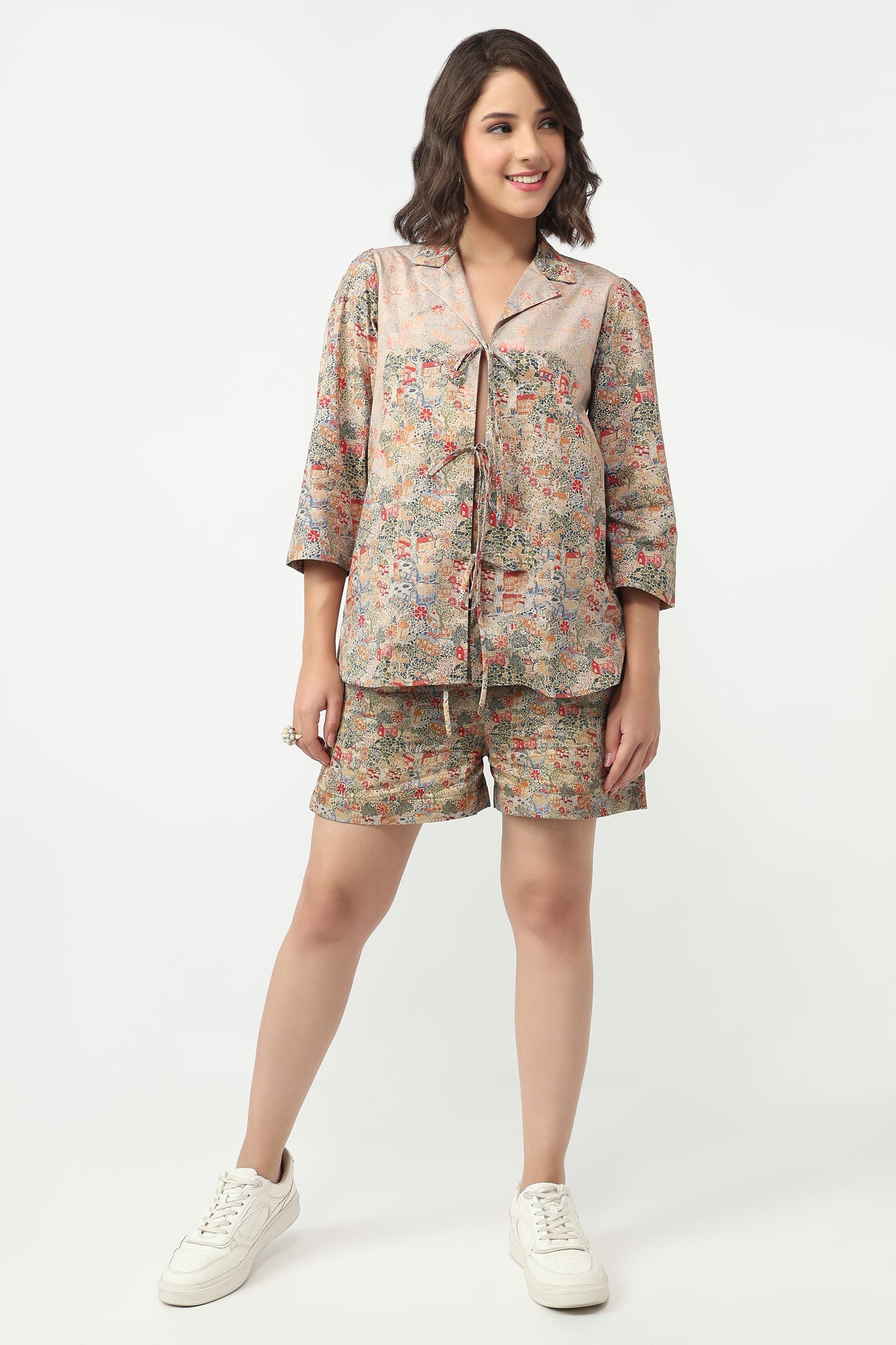 A woman modeling a matching two-piece pajama set featuring a short-sleeve button-up top and shorts in a beige base with a multicolor floral pattern, paired with white sneakers, against a plain white studio background.