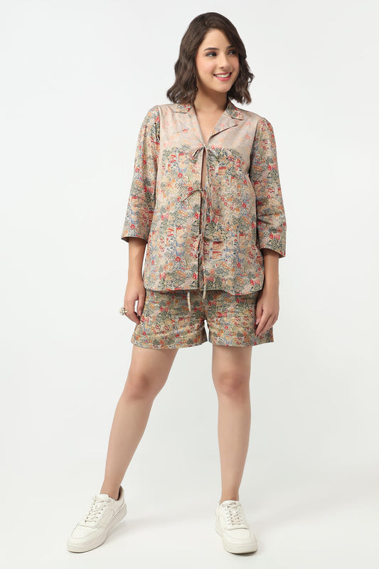 A woman modeling a matching two-piece pajama set featuring a short-sleeve button-up top and shorts in a beige base with a multicolor floral pattern, paired with white sneakers, against a plain white studio background.