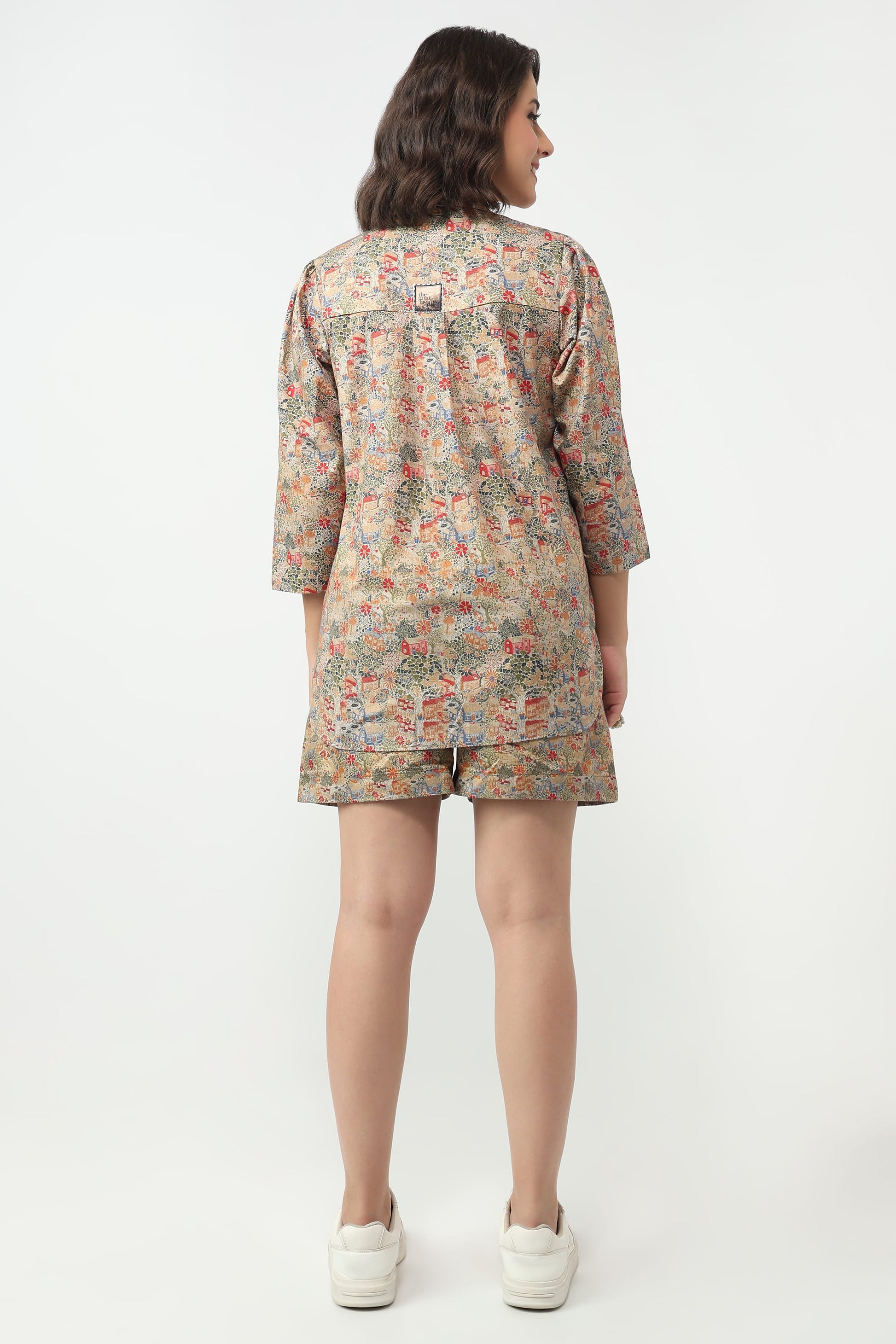A woman viewed from the back wearing a matching floral print pajama set consisting of a long-sleeve top and shorts, paired with white sneakers, against a plain white studio background.
