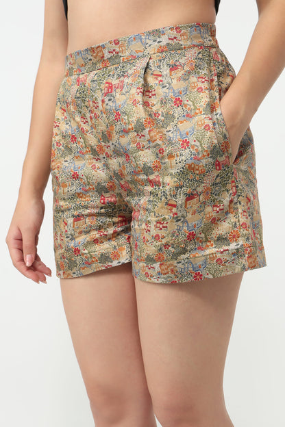 A close-up of a model wearing high-waisted shorts with a dense multicolor floral and botanical print on a beige base, set against a plain light gray background.