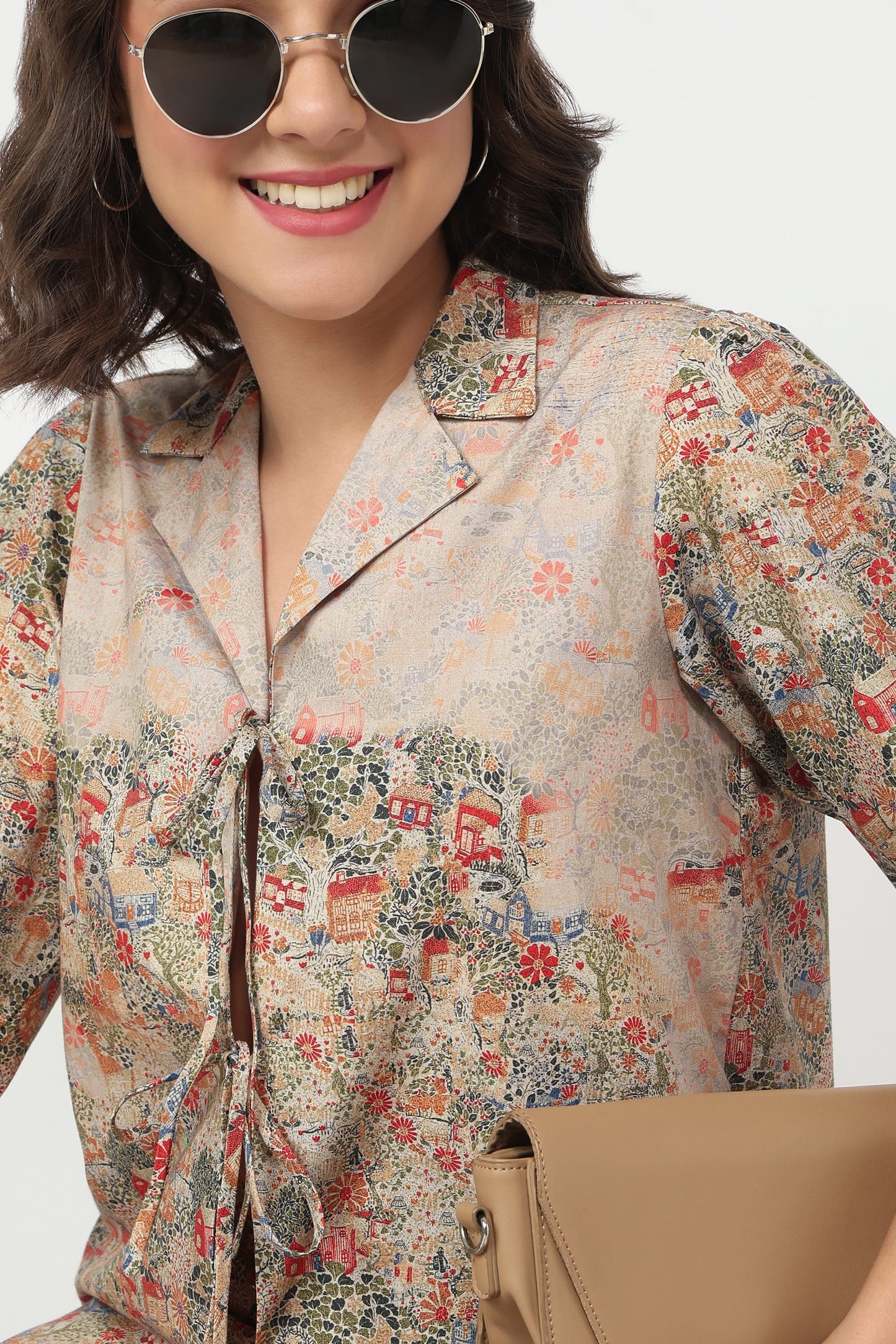 A woman wearing a beige short-sleeve button-up shirt with a colorful vintage-style illustrated print, styled with round sunglasses and a tan clutch bag against a light gray background.
