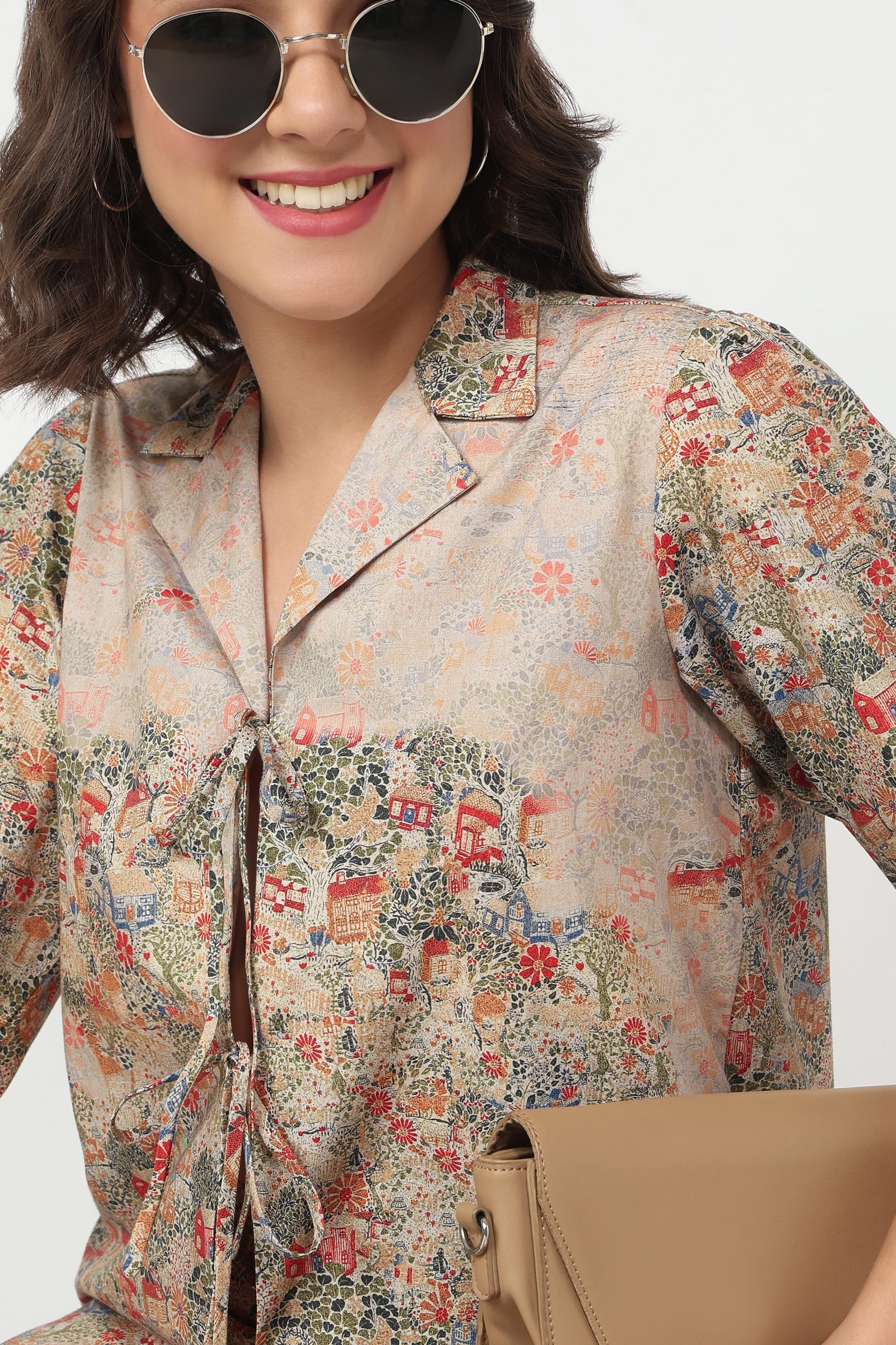 A woman wearing a beige short-sleeve button-up shirt with a colorful vintage-style illustrated print, styled with round sunglasses and a tan clutch bag against a light gray background.