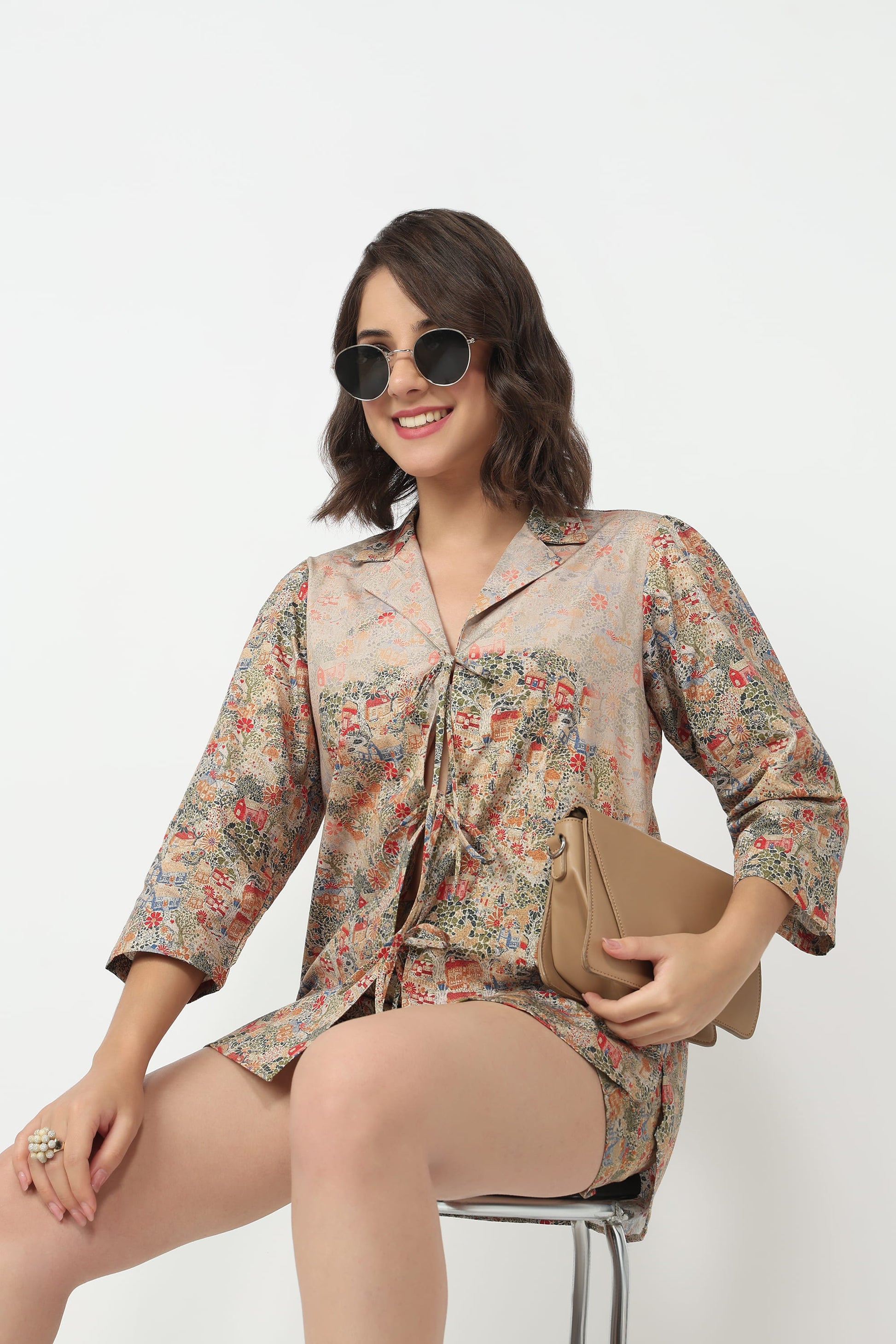 A woman wearing a beige button-up shirt dress with a multicolor floral print, sitting on a stool and holding a tan clutch bag.