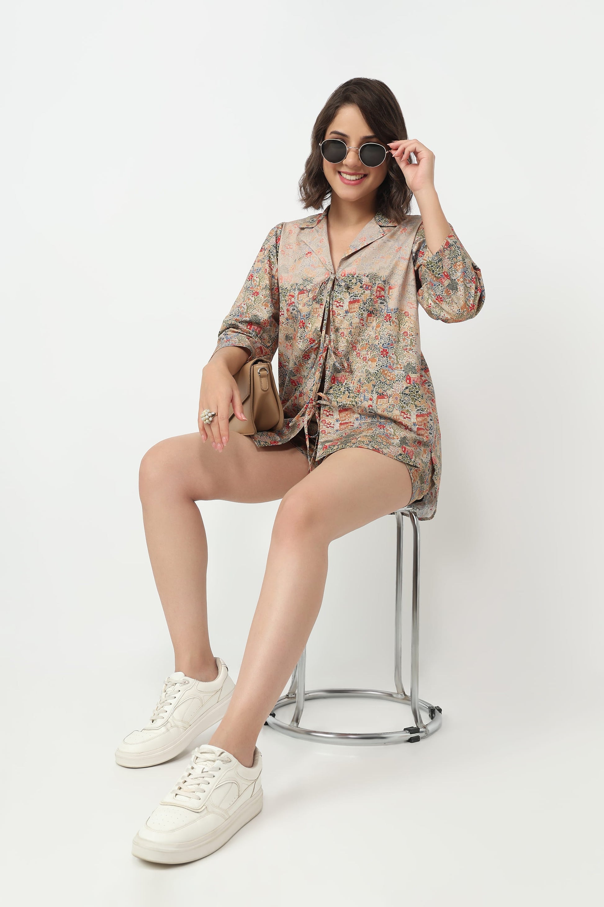 A woman sits on a metal stool wearing a short-sleeve button-front shirt dress with a beige and multicolor floral print, white sneakers, and sunglasses against a plain white studio background.
