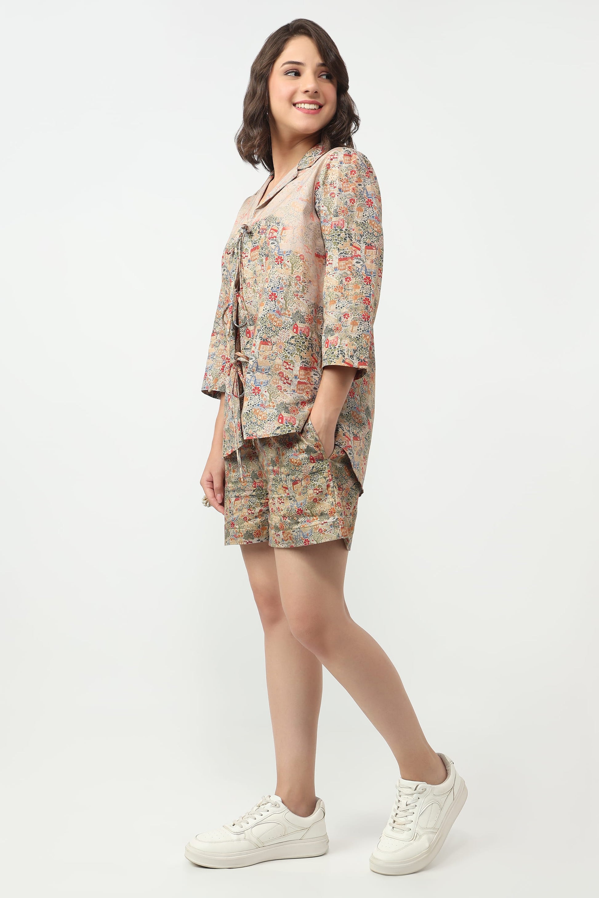 A woman modeling a matching two-piece floral print set consisting of a short-sleeve button-up shirt and shorts, paired with white sneakers, against a plain white studio background.