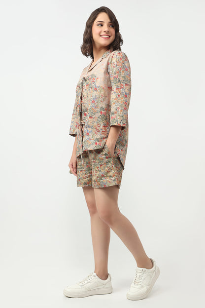 A woman modeling a matching two-piece floral print set consisting of a short-sleeve button-up shirt and shorts, paired with white sneakers, against a plain white studio background.