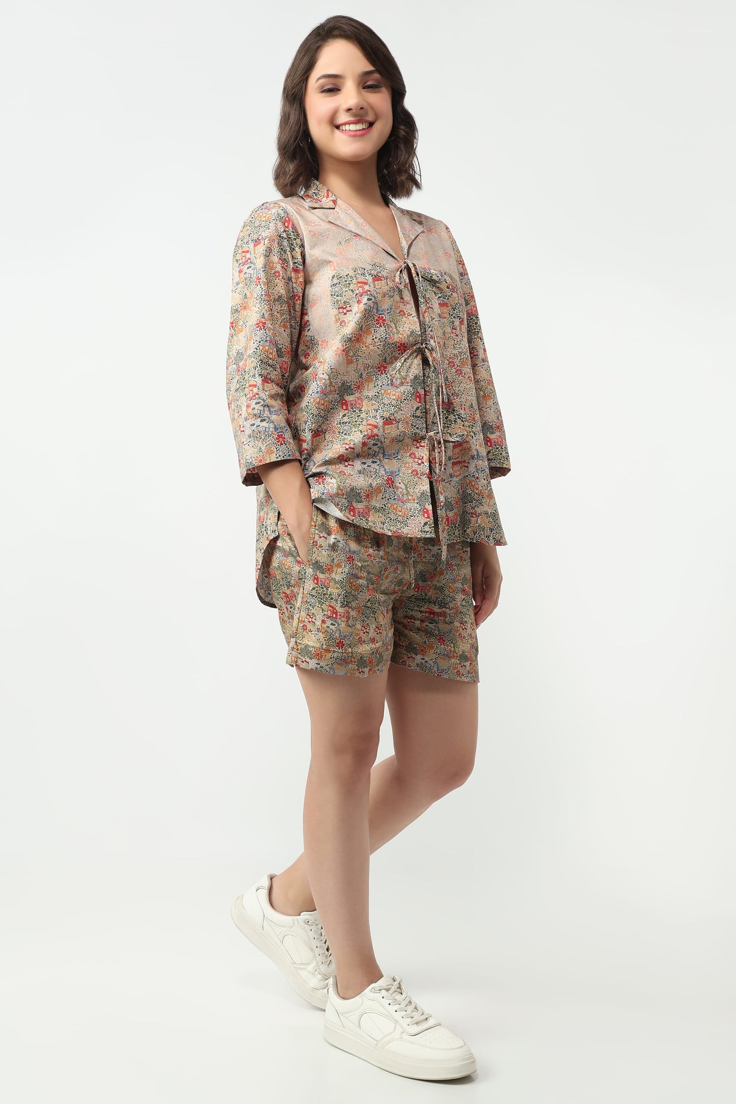 A woman modeling a matching two-piece pajama set with a long-sleeve button-up shirt and shorts, featuring a beige base with a multicolor floral pattern, paired with white sneakers against a plain white background.