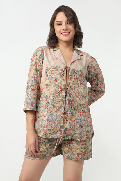 A woman modeling a two-piece pajama set featuring a long-sleeve button-up top and matching shorts, both in a beige base fabric with a dense multicolor floral print.