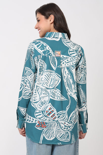 A woman shown from the back wearing a teal button-up shirt with a large white tropical leaf pattern and light blue jeans, against a plain light gray studio background.