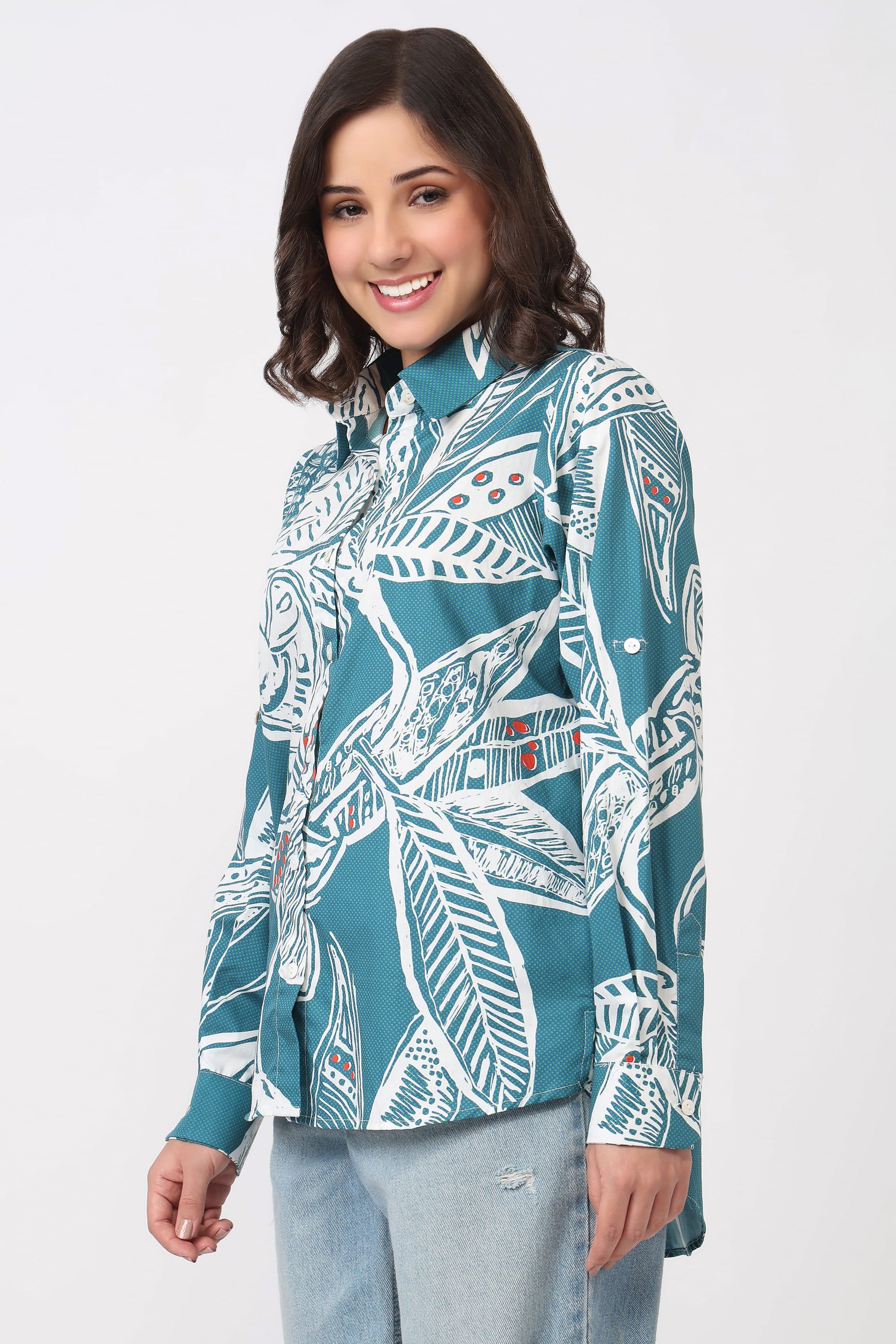 A woman wearing a teal and white tropical leaf print long-sleeve shirt paired with light-wash jeans, photographed against a plain light gray studio background.