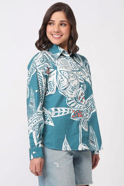 A woman wearing a teal button-down shirt with a white tribal-style graphic print and light-wash denim shorts, standing against a plain light gray studio background.