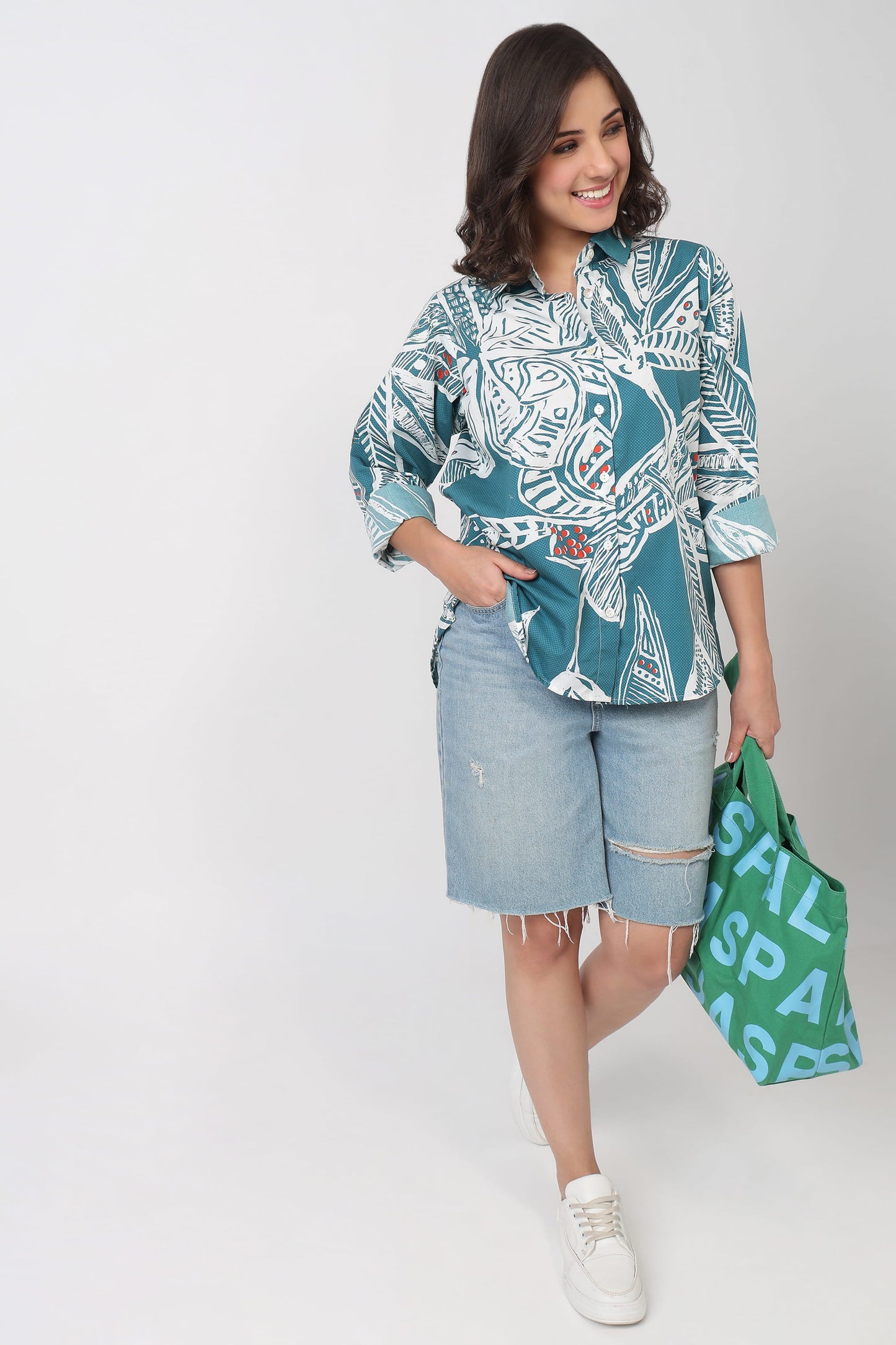 A woman modeling a teal and white patterned button-up shirt with light-wash denim shorts, holding a green shopping bag with white text against a plain light gray studio background.