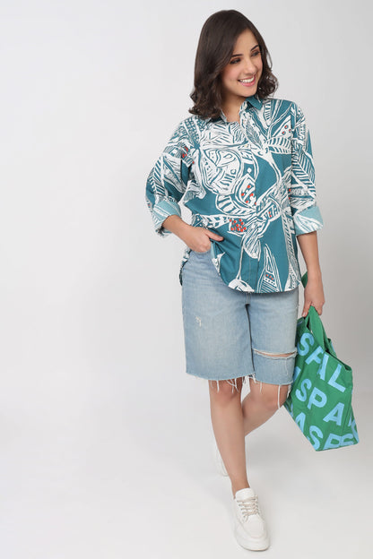 A woman modeling a teal and white patterned button-up shirt with light-wash denim shorts, holding a green shopping bag with white text against a plain light gray studio background.