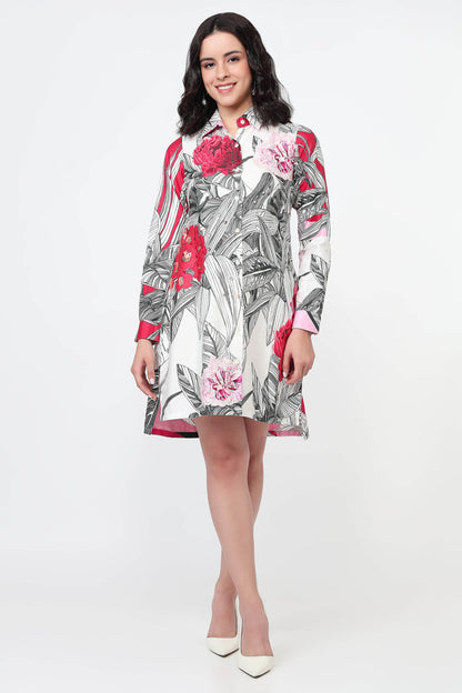 A woman modeling a long-sleeve mini dress with a white base and a bold floral print featuring red, pink, and gray botanical elements, paired with white pointed-toe heels against a plain white studio background.
