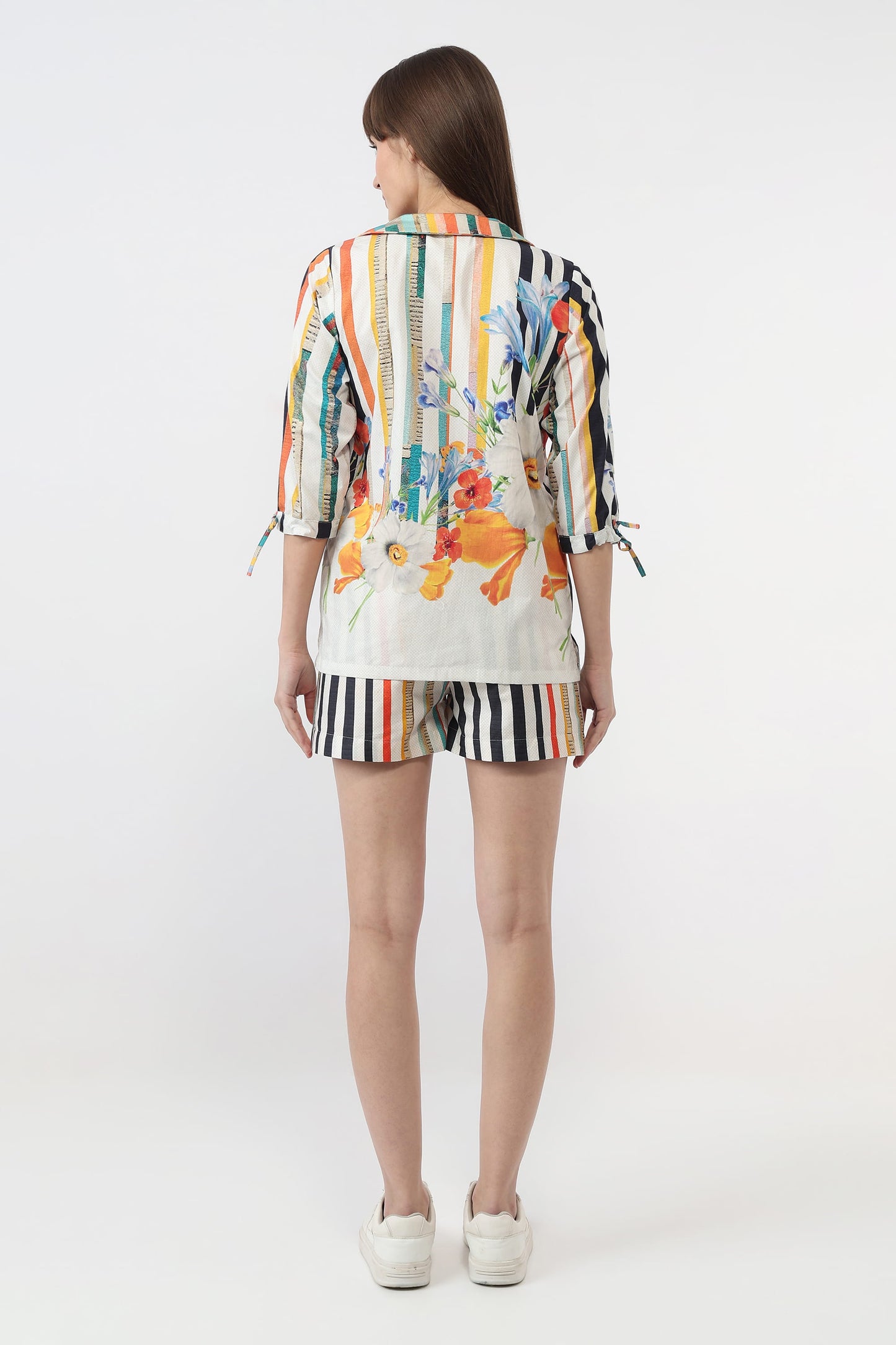 A woman viewed from the back wearing a colorful striped and floral print short-sleeve top with tie details at the sleeves, matching striped shorts, and white sneakers, against a plain light gray studio background.