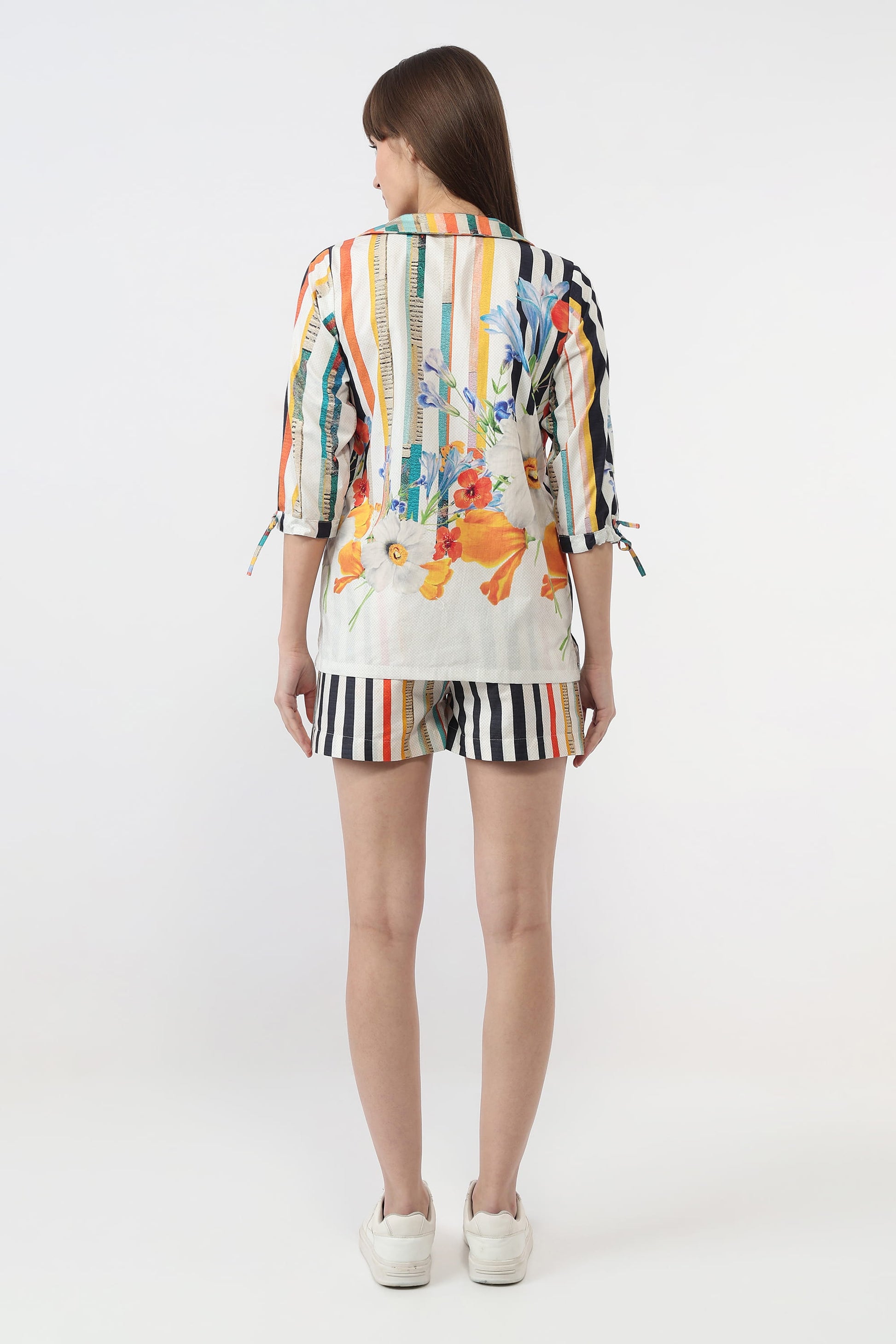 A woman viewed from the back wearing a colorful striped and floral print short-sleeve top with tie details at the sleeves, matching striped shorts, and white sneakers, against a plain light gray studio background.