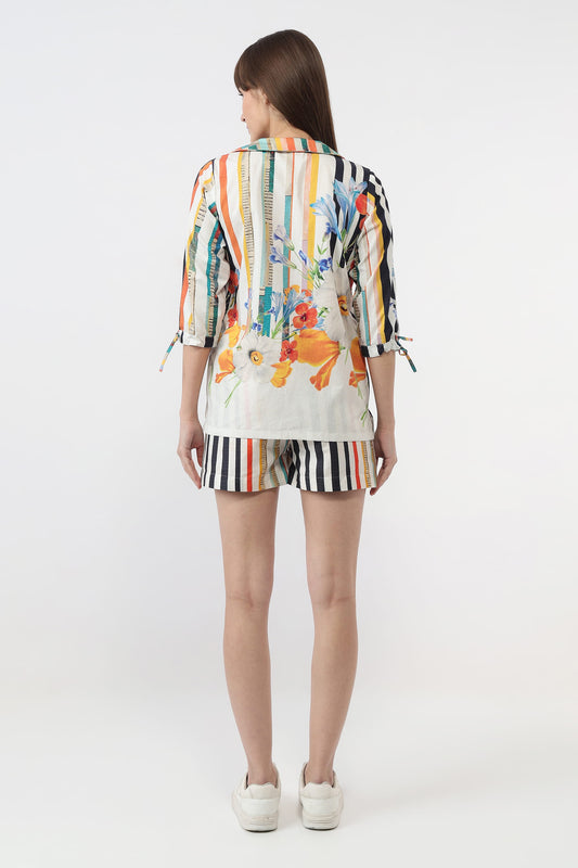 A woman viewed from the back wearing a colorful striped and floral print short-sleeve top with tie details at the sleeves, matching striped shorts, and white sneakers, against a plain light gray studio background.