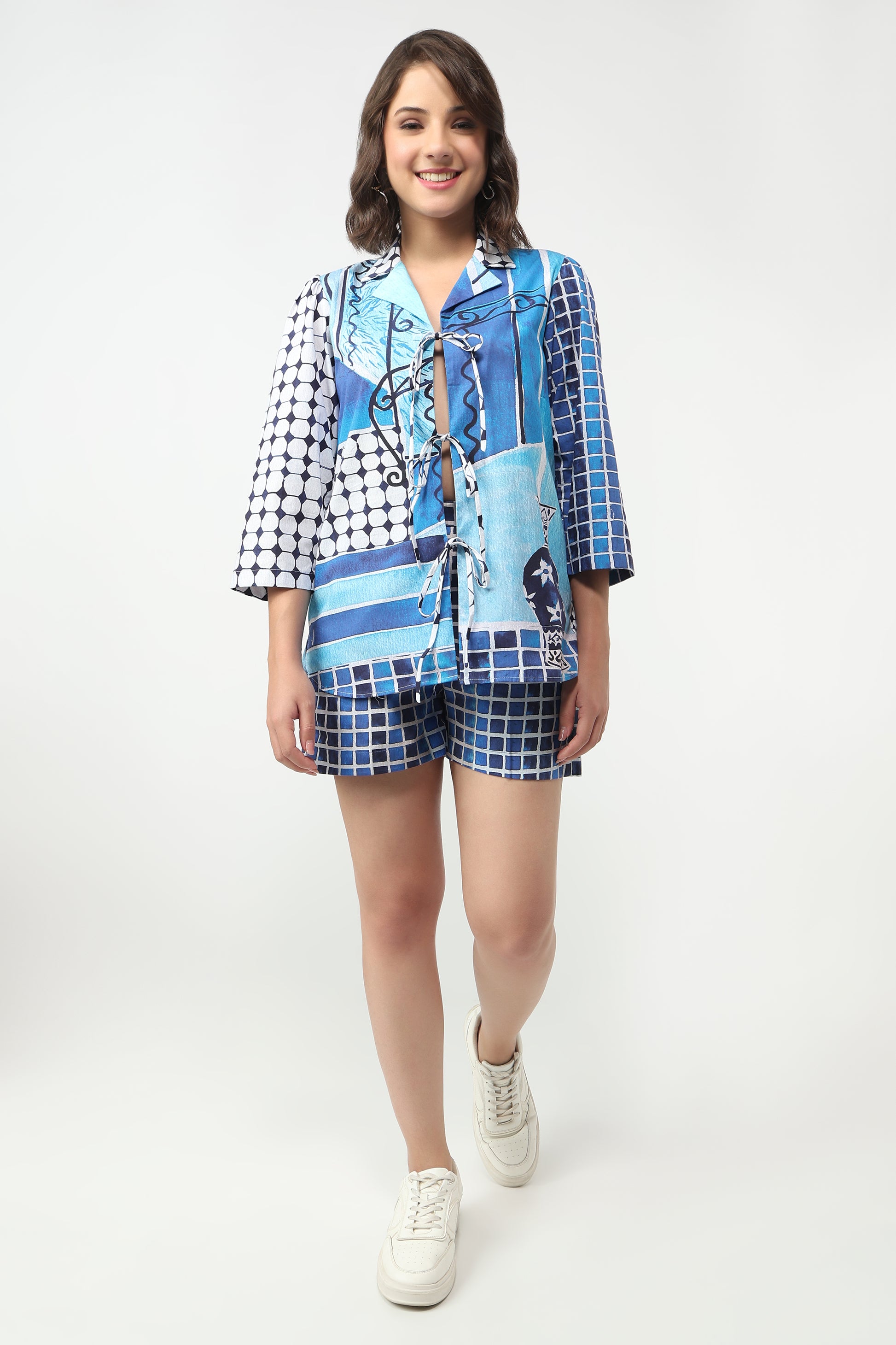 A woman modeling a matching two-piece lounge set featuring a blue and white patchwork print shirt and shorts, styled with white sneakers against a plain white studio background.