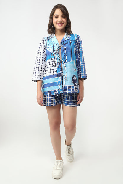 A woman modeling a matching two-piece lounge set featuring a blue and white patchwork print shirt and shorts, styled with white sneakers against a plain white studio background.