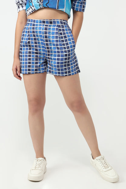 A model wearing blue and white checkered grid-patterned shorts, a matching blue patterned crop top, and white sneakers, shown from the waist down against a plain white studio background.