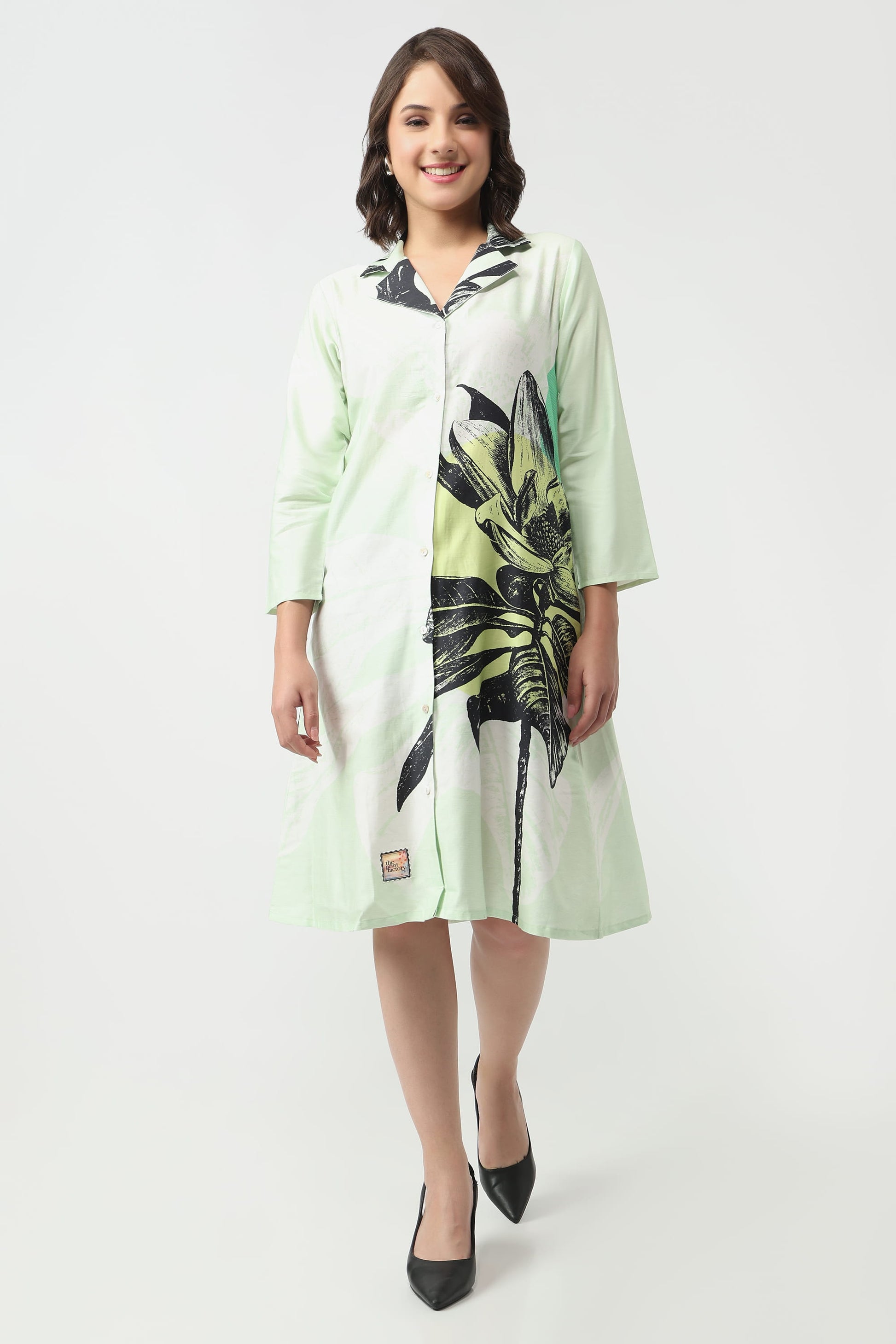 A woman wearing a light mint green knee-length shirt dress with a black and green tropical leaf print on the front panel, styled with black pointed-toe heels against a plain light grey studio background.