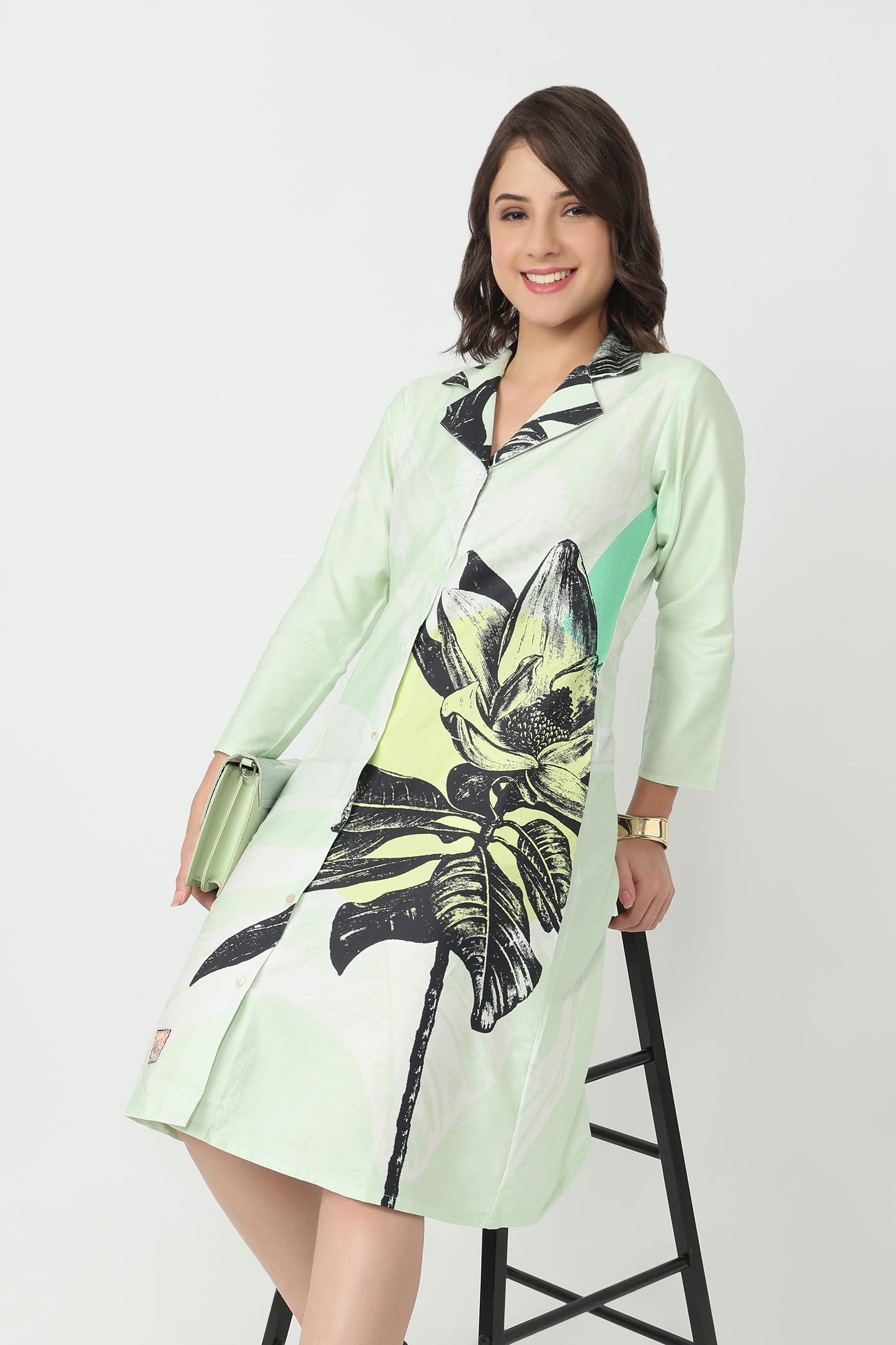 A woman wearing a mint green long-sleeve shirt dress with a black and yellow tropical leaf print, standing on a black ladder against a white studio background.